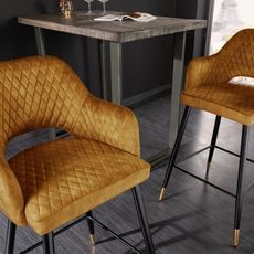 Estila Art deco zamatová barová stolička Fribourg v horčicovo žltej farbe s opierkami na čiernych nožičkách 104cm